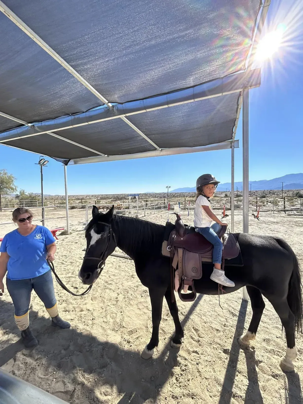 A young girl in a riding helmet sitting on a horse while an instructor leads it under a shade structure